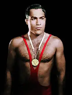 Muskulöser junger Mann im roten Ringersportsdress und kaum bedachten Oberkörper mit einer goldenen Medaille an einer weißen Kordel um den Hals von vorn und schwarzem Hintergrund in Farbe.