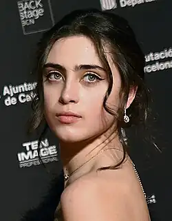 Maria Morera (2022) mit langen, dunklen Haaren und auffälligem Make-up und schaut über die Schulter. Sie trägt ein schwarzes elegantes Outfit und eine Halskette dazu kombiniert. Die Frisur ist dunkel und hochgesteckt, wobei ein hängender Ohrring sichtbar ist. Der Hintergrund des Bildes ist eine schwarze Wand mit mehreren weißen Schriftzügen. Dazu gehören „BACK stage BCN“, „Ajuntament de Barcelona“, „Diputació de Barcelona“ und „TEN IMAGE PROFESSIONAL“.