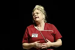 Marie Schöneburg in ihrer Rolle als Nancy im Schlosspark Theater (2023). Sie trägt eine rote Uniform mit weißen Akzenten, die ein Namensschild mit der Aufschrift „Nancy“ sowie Symbole für Barrierefreiheit enthält. Ihr blondes Haar ist hochgebunden, und sie hält ihre Hände vor ihrem Körper zusammen. Sie hat einen nachdenklichen Gesichtsausdruck. Der Hintergrund ist schwarz.