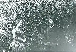 Schwarz-Weiß-Fotografie von zwei Personen vor einem Gitter mit Kletterpflanze. Die Frau auf der linken Seite trägt ein Kleid und hochgesteckte Haare. Der Mann auf der rechten Seite Vollbart und einen Anzug. Sie sind einander zugewandt, halten aber Abstand. Die Frau lächelt.