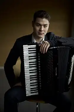 Marius Staible sitzt im Anzug auf einem Stuhl und hält mit der linken Hand ein Akkordeon auf dem Oberschenkel.