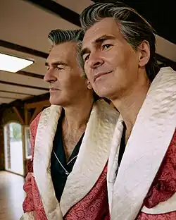 Mark Scheibe with neatly styled, gray-streaked hair wears a red patterned robe with a quilted white collar. He gazes into a mirror, creating the impression of two identical figures standing side by side in a warmly lit interior.