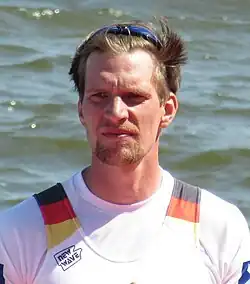 Maximilian Musnki steht bei der Siegerehrung im Nationaltrikot vor der Regattastrecke. Seine Sonnenbrille hat er in die Haare hochgeschoben.