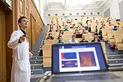 Ein Dozent in einem weißen Kittel steht vor den mit Studierenden gefüllten Sitzreihen des Hörsaals 1.