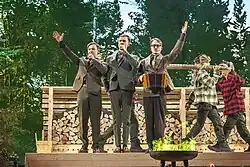 Die finnlandschwedischen Band KAJ auf dem Melodifestivalen 2025 in Malmö auf einer Bühne, die vor einem Hintergrund aus Bäumen steht. Im Vordergrund befinden sich drei Gruppenmitglieder, die Anzüge tragen. Zwei von ihnen halten Mikrofone, und eine Person spielt ein Akkordeon. Dahinter sind zwei Personen in karierten Hemden und Hüten zu sehen, die ein großes Holzstück tragen. Im Hintergrund gibt es einen Stapel gehacktes Holz und eine Holzstruktur. Im Vordergrund ist eine Feuerstelle mit grünen Flammen sichtbar. Die Bühne scheint eine in einem Wald-ähnlichen Umfeld zu stehen.