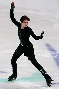 Michail Schaidorow steht auf der Eisfläche in einer kampfsportähnlichen Pose. Er trägt einen schwarzen Anzug mit silbernen Schnallen und senkrechten grünen Streifen, der an die Kleidung der Hauptfiguren des Films „Matrix“ angelehnt ist.