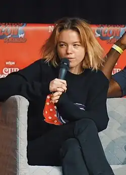 Milly Alcock mit unordentlichem, blondem Haar, auf einem grauen Sessel sitzt und ein Mikrofon in der Hand hält. Sie trägt trägt einen schwarzen Pullover mit einem bunten Muster darauf sowie schwarze Hosen. Milly Alcock hat einen nachdenklichen Gesichtsausdruck. Im Hintergrund ist ein rotes Banner zu sehen, auf dem der Text „Comic Con Dortmund“ geschrieben steht. Ebenfalls sind Logos von „Nissin“ und „Cup Noodles“ auf dem Banner abgebildet. Neben der Person ist der Arm einer weiteren Person sichtbar, die ebenfalls auf einem Sessel sitzt. Das Foto wurde auf der Comic Con in Dortmund 2022 aufgenommen.