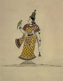 Frauengestalt jüngeren Alters, barfuß, sie trägt ein gelbes Kleid mit einem Muster aus roten Blumen und ein rotes Oberteil, sie blickt nach links auf den grünen Vogel, der auf ihrer rechten Hand sitzt. Auf dem Kopf trägt sie eine hohe goldene Haube