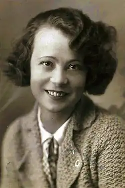 Debelak Deržaj blickt frontal lächelnd in die Kamera, sie hat gewelltes, dunkelblondes Haar und trägt über einer hellen Bluse eine Strickjacke.