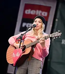 Miss Allie singt auf der Bühne des Rockspektakel in Hamburg 2018 und spielt eine Akustikgitarre. Sie hat blondes Haar und trägt einen rosa Pullover, eine graue Hose. und eine schwarze Mütze. An der Gitarre ist ein auffälliges rotes Schild angebracht, auf dem „Miss Allie“ steht. Im Hintergrund ist ein Banner mit der Aufschrift „delta radio“ zu sehen.