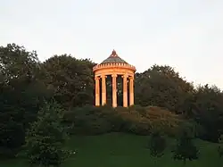 Farbfotografie in der Untersicht eines Rundtempels mit Säulen und einer goldenen Kugel als Spitze. Der Tempel steht auf einem Hügel mit Büschen und ist von Bäumen umgeben.