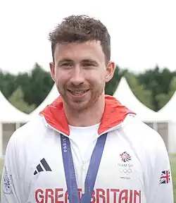 Morgan Bolding trägt eine weiße Jacke mit der Aufschrift Great Britain. Um den Hals trägt er ein blaues Band, an dem seine Goldmedaille befestigt ist, die aber nicht im Bild ist.
