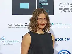 Morgane Ferru mit lockigen Haaren, bei der Verleihung der Deutschen Akademie für Fernsehen 2015, trägt ein ärmelloses schwarzes Kleid und lächelt. Sie steht vor einem Hintergrund mit verschiedenen Logos und Texten. Der Hintergrund enthält folgende Texte und Logos: „DEUTSCHE AKADEMIE FÜR FERNSEHEN“, „Minister für Bund, Europa und Medien des Landes Nordrhein-Westfalen und Chef der Staatskanzlei“, „CROSS X CHECK high speed camera systems“, „pensionskasse rundfunk“, „producer“. Ein Logo mit den Buchstaben „V“ und „W“, die sich überlappen.Weitere Logos, die teilweise sichtbar, aber nicht vollständig lesbar sind.