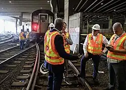 Blick aus dem Gleisbett, ein Dutzend Männer in MTA-Warnwesten arbeiten oder debattieren vor einem abgestellten U-Bahn-Zug.