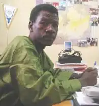 Moussa Kaka, ein afrikanischer Mann mit kurzem Haar, sitzend an einem Tisch mit Stift in der Hand.