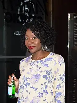 Muna Otaru mit lockigem, schwarzem Haar bei den Toronto International Film Festival 2014, die vor einer Glastür oder einem Fenster steht. Sie trägt ein langärmeliges Hemd mit einem floralen Muster in Lila- und Weißtönen. In der linken Hand hält sie eine grüne Glasflasche mit einem Etikett und einem Strohhalm. Im Hintergrund ist auf der Glasscheibe Schrift zu erkennen, darunter das Wort „HOLLY“ sowie andere, teilweise verdeckte Wörter. Auf der rechten Seite des Bildes befindet sich ein vertikales Schild mit Text, der verschiedene Artikel oder Dienstleistungen auflistet.