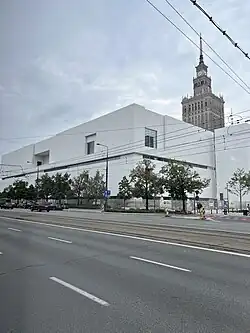 Ein weißer Flachbau steht vor dem Turm des Kulturpalastes in Warschau.