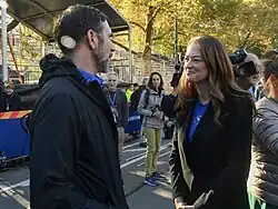 Aufnahme in einem herbstlichen Stadtpark. Zwei Personen groß im Bild: Laura Kavanagh, rechts, im Gespräch mit Jamie Torres-Springer, Abteilungsleiter der Metropolitan Transportation Authority, links. Daneben und im Hintergrund einzelne Zuschauer und Pressevertreter.