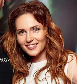 Natalka Denysenko (2023), mit langen, lockigen, rotbraunen Haaren. Sie trägt ein schlichtes, weißes Oberteil, das bis zum Hals reicht und keinen besonderen Schmuck aufweist. Neben der Person ist der Oberkörper eines anderen Menschen zu sehen, der ein schwarzes Kleidungsstück trägt, auf dem weiße Buchstaben oder Symbole zu erkennen sind. Der Hintergrund des Bildes ist verschwommen und überwiegend grünlich mit einigen dunkleren Bereichen, die an eine Naturumgebung oder einen parkähnlichen Ort erinnern.