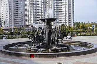 Ein runder Brunnen mit verschiedenen Figuren in der Mitte. Aus einer Art Schale die erhoben dargestellt ist, sprudelt Wasser.