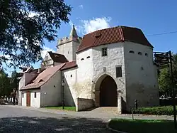 Mittelalterliches Tor, weiß verputzt, Dachziegel oben, links davon ein Türmchen mit Zinnen und einer steinernen Spitze in der Mitte