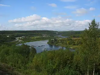 Foto eines Flusses, der durch eine grüne Landschaft fließt. Am Fluss eine kleinere Siedlung.