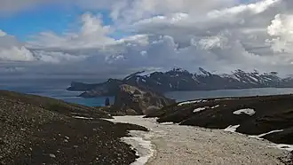 Blick über die Landspitze Fildes Point mit dem Brandungspfeiler Petes Pillar in der Meerenge Neptunes Bellows hinweg auf Mount&nbsp;Kirkwood (Hintergrund)
