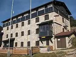 Blick auf zwei Seiten einer großen, aus groben Steinen gemauerten Berghütte, der Neuen Traunsteiner Hütte; davor zwei leere Fahnenmasten