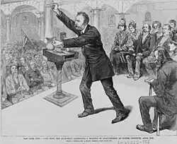 Most steht mit erhobenener Faust und einem Krug in der anderen Hand am Rednerpult und spricht in den gut gefüllten Saal.