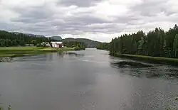 Foto eines breiteren Flusses in bewaldeter Landschaft, am Ufer links im Bild vereinzelte Häuser