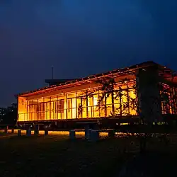 Ein beleuchtetes, einstöckiges Gebäude mit Glasfassade in der Nacht. Vor diesem befinden sich auf einer Grasfläche Picknickbänke.