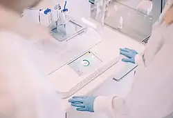 Eine Person im weißen Kittel und mit blauen Gummihandschuhen steht vor einem Labortisch, auf dem ein 3D-Bioprinter steht. Dieser besteht aus einer vertikal ausgerichteten Spritze, Linearantrieben und einer Petrischale