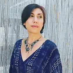 Porträt einer Frau mit langen, zurückgebundenen Haaren, sie steht seitlich und hat den Kopf der Kamera zugewandt. Sie trägt ein blau-weiß gemustertes Oberteil und eine aufwändige Silberkette