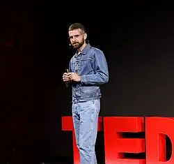 Olaf Grawert steht auf der Bühne eines TED Talks, trägt eine Jeansjacke und Jeanshose, spricht ins Mikrofon und hält ein Objekt in den Händen. Im Hintergrund ist das rote TED-Logo teilweise sichtbar.