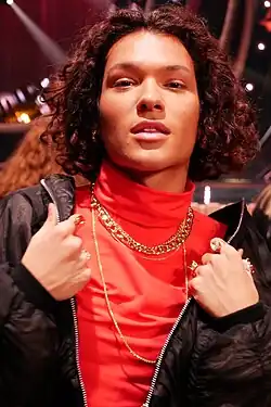 Omar Rudberg auf dem Melodifestivalen 2019, mit lockigem Haar, der ein auffälliges rotes Oberteil trägt, kombiniert mit einer schwarzen Jacke. Er hat mehrere goldene Halsketten um den Hals, die dem Gesamtbild einen Hauch von Eleganz verleihen, und er trägt zahlreiche Ringe an den Fingern, die alle individuell gestaltet sind. Die Hände der Person sind in einer Bewegung, als ob sie die Jacke zurechtrückt oder präsentiert.Der Hintergrund ist unscharf gehalten, aber es lassen sich Lichter erkennen, die auf eine Bühne oder einen Veranstaltungsort hinweisen könnten. Diese Lichter verleihen dem Bild eine besondere Atmosphäre.Der Fokus liegt auf der modischen Darstellung und den prägnanten Details der Accessoires, die den Stil von Omar Rudberg unterstreichen.