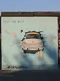 Frontale Farbfotografie von der Berliner Mauer mit einem Bild von einem weißen Auto, dem Trabant, der eine hellblaue Wand in Richtung Betrachter durchbricht. Links oben steht „Test the best“.