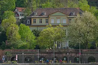 Ein lindgrünes Gebäude, vorne verdeckt von frühlingshaft grünen Bäumen im Garten vor dem Gebäude auf der großen Sandsteinterasse.