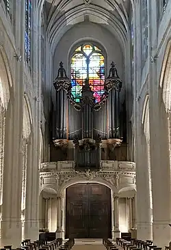 Das Foto zeigt den Blick aus einiger Entfernung auf die Hauptorgel und die zugehörige kleine Brüstungsorgel auf der Empore der Kirche Saint-Gervais. Hinter der Orgel sieht man Teile eines hohen bunten Kirchenfensters, das in den Farben Gelb, Grün, Rot, Blau und Weiß im Gegenlicht leuchtet; im Vordergrund des Fotos sieht man hohe weiße Pfeiler und einige Reihen mit dunklen hölzernen Sitzhockern.