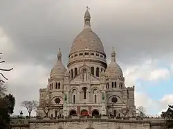 Frontale Farbfotografie in der Untersicht eines weißen Kuppelbaus im neobyzantinischen Stil. Es gibt eine mittlere, große Kuppel und zwei kleinere Kuppeln an beiden Seiten. Alle Kuppeln haben an der Spitze kleine Säulenrundgänge mit Kreuzen und an der Basis breite Säulenrundgänge. Der mittlere Vorbau besteht aus einem Dachgiebel, Rundbogenfenstern und Rundbogeneingängen, mehreren Steinfiguren und zwei grünen Reiterstatuen auf Sockeln.