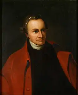 Ein sitzender Patrick Henry mit einem schwarzen Anzug und einer roten Jacke vor dunklem Hintergrund.