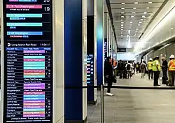 Übersicht einer Bahnhofshalle der Penn Station mit Fahrgästen und Arbeitern in Warnwesten. Links am Bildrand gut lesbar ein hoher Informations-Bildschirm. Unter Überschrift MTA Long Island Rail Road und der Uhrzeit 11:04:01 zeigt er eine alphabetische Liste von Zielbahnhöfen beginnend bei A wie „Albertson“ bis D wie „Deer Park“. Hinter jedem Stationsnamen steht die Abfahrtszeit des nächsten Zugs und der Linienname auf dem Hintergrund eines rechteckigen, in Linienkennfarbe codierten Feldes. Am Ende vieler Zeilen steht die Nummer des Abfahrtgleises in Penn Station.