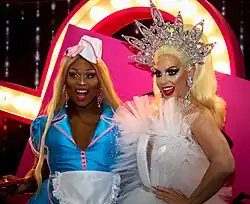 Porträtfoto einer vor einem rosa Hintergrund sitzenden Peppermint. Sie trägt eine hellblaue Kellnerinnenuniform mit rosa Umrandungen, eine weiße Schürze und einen kleinen, oben leicht spitzen und unten flachen, weißen Hut mit roten Streifen. Daneben trägt sie silberne Ohrhänger an beiden Ohren, rosa Lippenstift, Wimperntusche, rosa Lidschatten und eine blonde, bis zur Brust reichende Perücke. In der rechten Hand hält sie ein rotes Serviertablett. Links neben ihr sitzt ihre Kollegin Alyssa Edwards, die ein weißes Federnkleid, eine blonde, nach oben stehende Perücke und ein silbernes Diadem mit mehreren Spitzen trägt. Hinter ihnen ist ein roter Bogen mit mehreren Glühbirnen zu sehen.