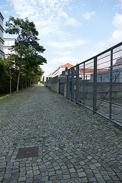 Mit Kopfsteinengepflasterter moderner Weg, links von Mehrfamilienhäusern und Bäumen gesäumt, rechts von einem Metallzaun