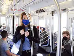 Feinberg zwischen Reihen von Haltestangen stehend in einem U-Bahn-Zug mit mehreren sitzenden Fahrgästen bei Tageslicht, Stoffbeutel mit Gesundheitsartikeln für die Fahrgäste über der Schulter, Mobiltelefon in der Hand