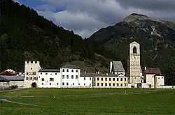Frontale Farbfotografie einer Klosteranlage mit Bergen im Hintergrund. Mehrere Häuser stehen auf einer Alm nebeneinander und rechts ist eine Turmuhr.