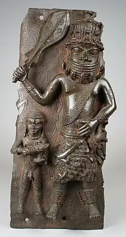 Zwei Figuren auf einer Reliefplatte, davon ist eine halb so groß wie die andere. Die größere Figur trägt einen Rock und einen Hut. Um seinen freien Oberkörper ist ein Kette aus mehreren Strängen geschlungen. In der rechten Hand hebt er ein Zeremonialschwert mit breiter Klinge nach oben. Die kleiner Figur ist nackt und am ganzen Körper tätowiert.