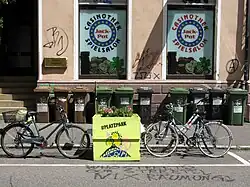 Zwei Fahrräder stehen beidseitig in Fahrradständern eines bunt bemalten Hochbeetes, das auf der Markierung eines Pkw-Stellplätzes steht. Im Hintergrund die Fassade eines Ladengeschäfts