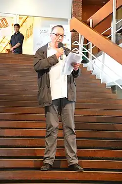 Ein Mann mit Brille steht auf einer Treppe, er hält ein Mikrofon und blickt auf ein Skript, das er in den Händen hält