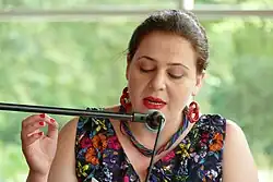 Foto einer Frau, die konzentriert nach unten blickt und in ein Mikrofon spricht. Man sieht ihren Oberkörper, sie trägt ein geblümtes Kleid und große rote Ohrringe, sowie eine bunte Kette..