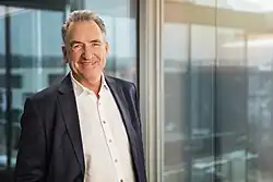 Ein Porträt von Joachim Lang. Er trägt ein weißes Hemd ohne Krawatte, darüber ein blaues Jackett, das offen steht. Er hat kurze graue Haare und Geheimratsecken. Im Hintergrund eine hohen Bürofensterfront.